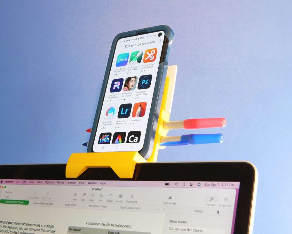 yellow 3D printed phone holder mounted on a laptop computer screen holding a phone and two pens